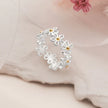 A silver daisy-patterned women silver ring with small gold centers, displayed on a soft beige fabric background with a pink flower in the corner.